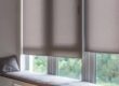 Motorized beige roller shades cover the top half of a window next to a windowsill nook.
