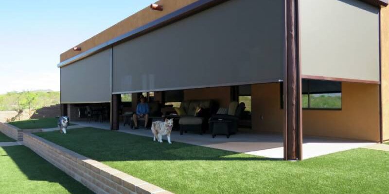 Solar screen halfway down across a patio opening with the homeowners relaxing inside and their two dogs run across the grassy yard.